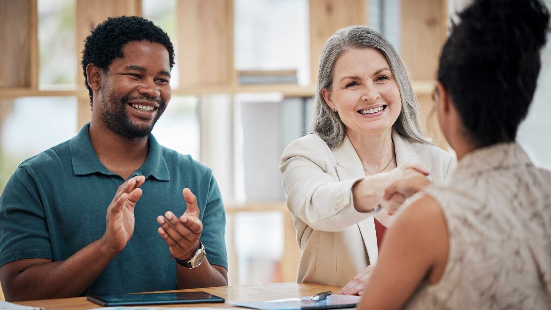 A woman and a man seated at a table, the woman shakes another woman's hand as she welcomes her to the team and the man claps