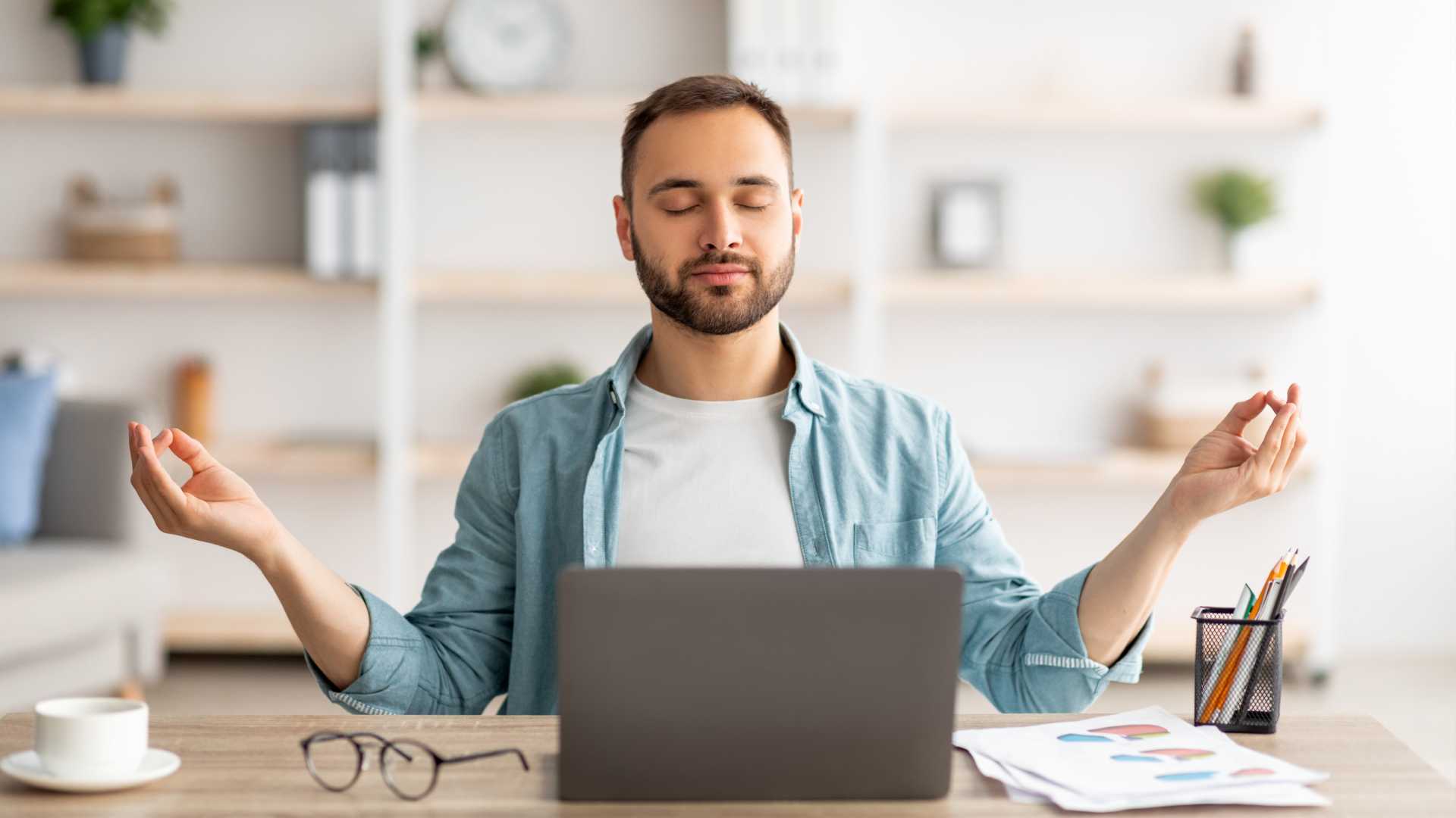 A casual businessman sits at his laptop in his home office; he is holding his hands up and to the sides in a meditative pose, and his eyes are closed. His face is serene. He is upskilling or possibly reskilling.