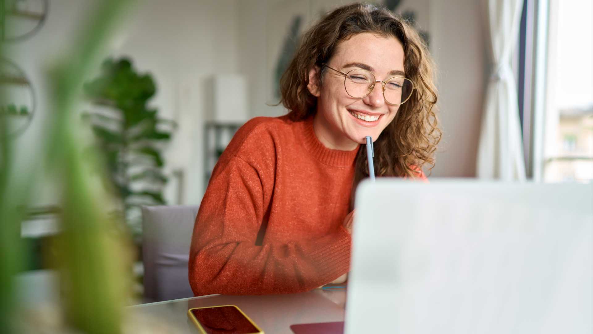 A smiling woman sits at a desk in front of an open laptop