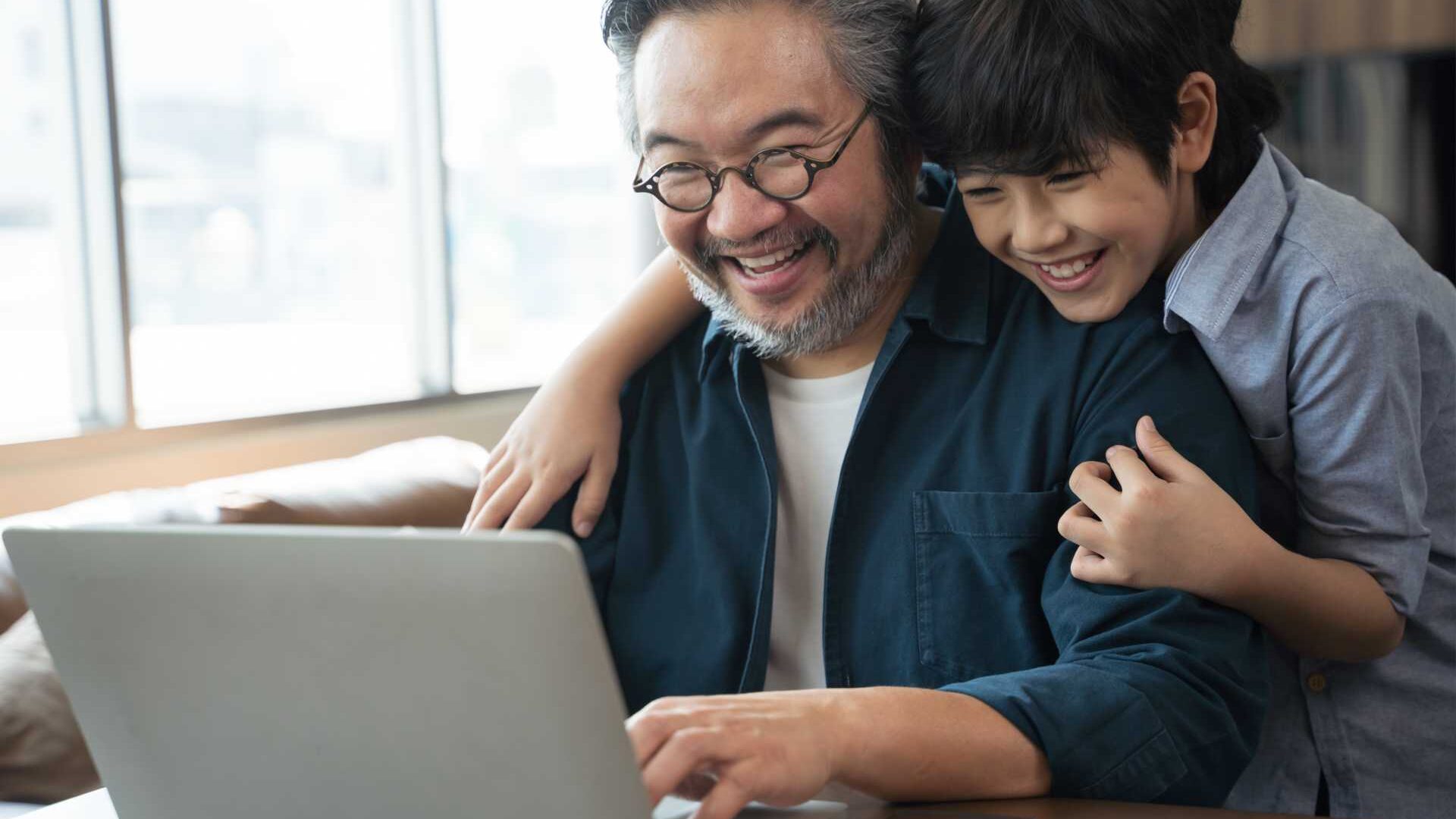 A man works from home in front of an open laptop; his child gives him a hug