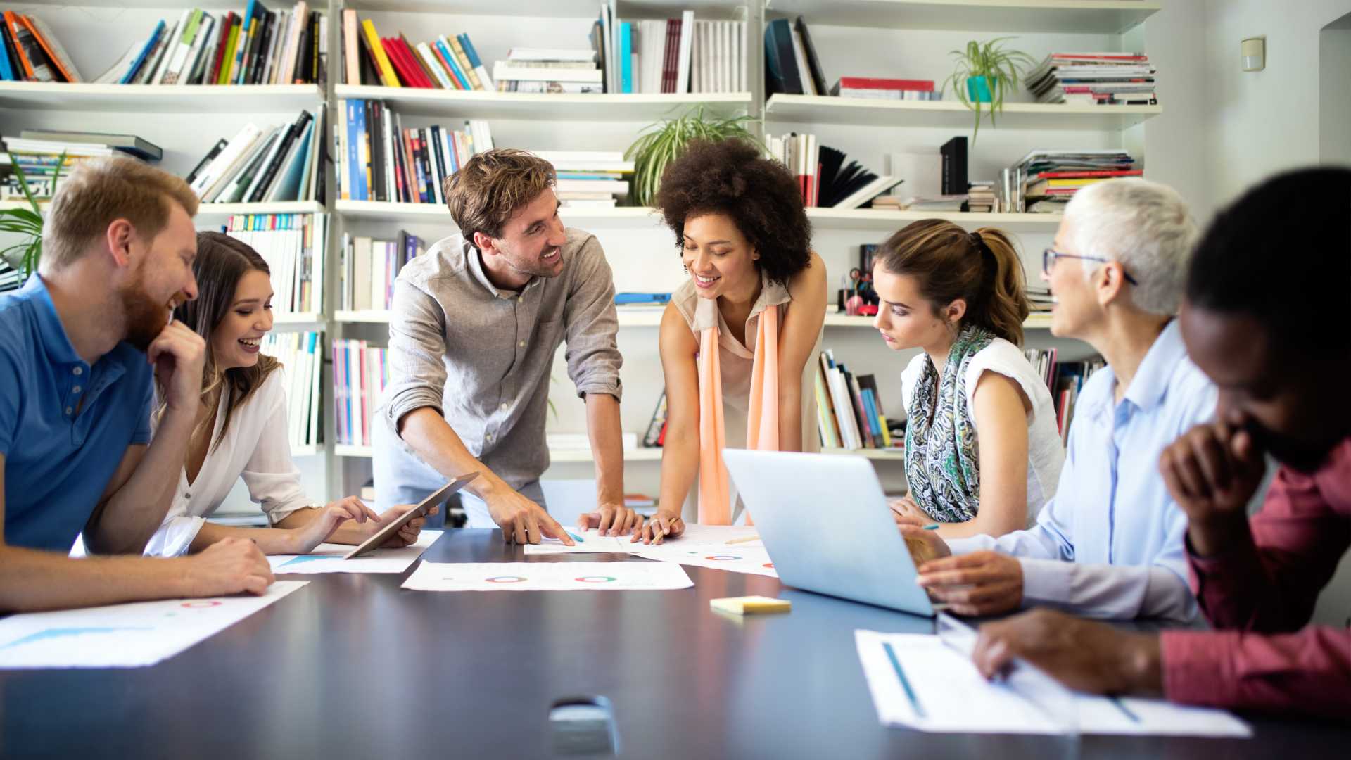 A diverse group of coworkers sits at a conference table in a brightly lit office space.