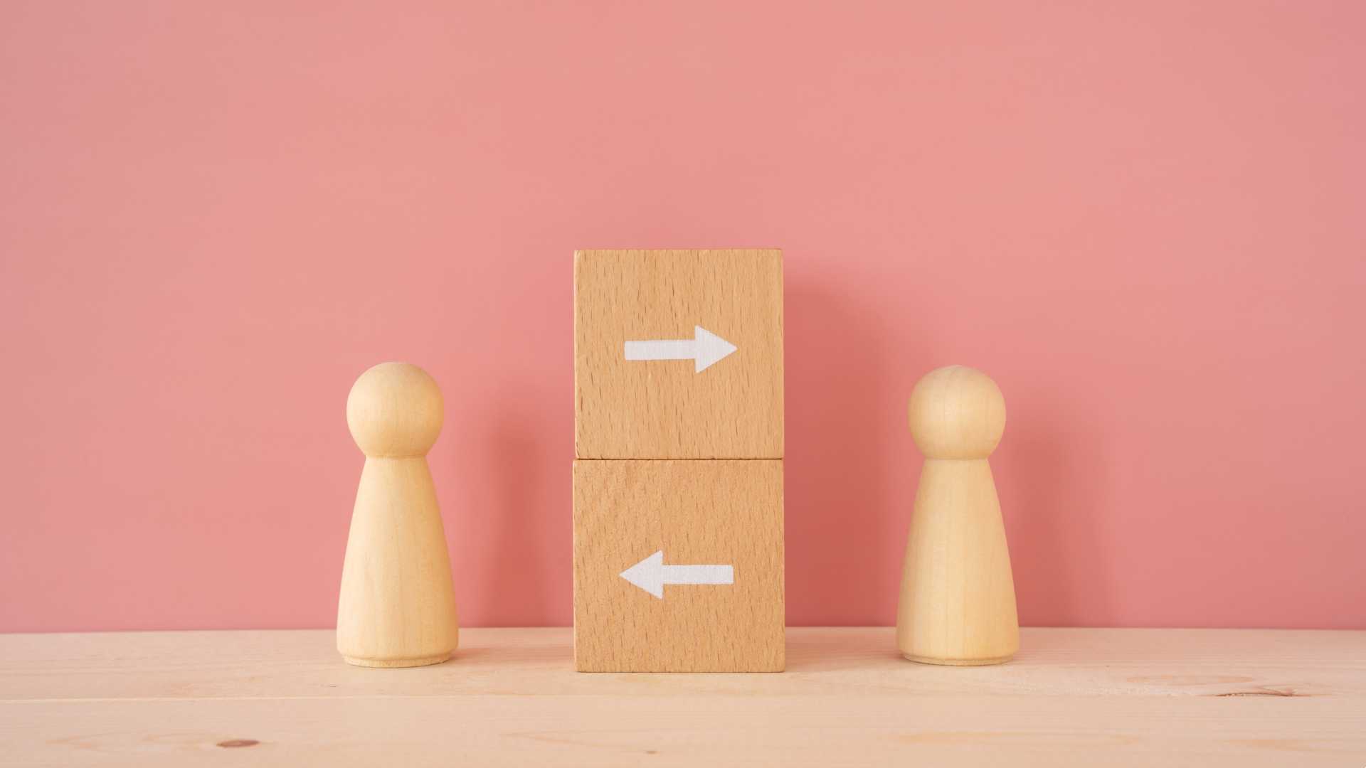 Two wooden block human figures standing against a pale pink background with two wooden blocks stacked up in the middle of them with painted white arrows going in either direction.