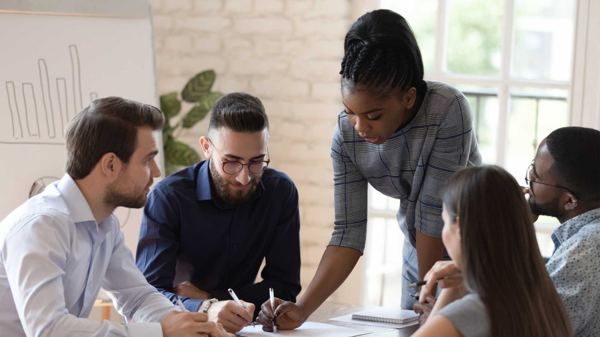 A group of coworkers are having a discussion while sitting at a table with a graph on the chart behind them.