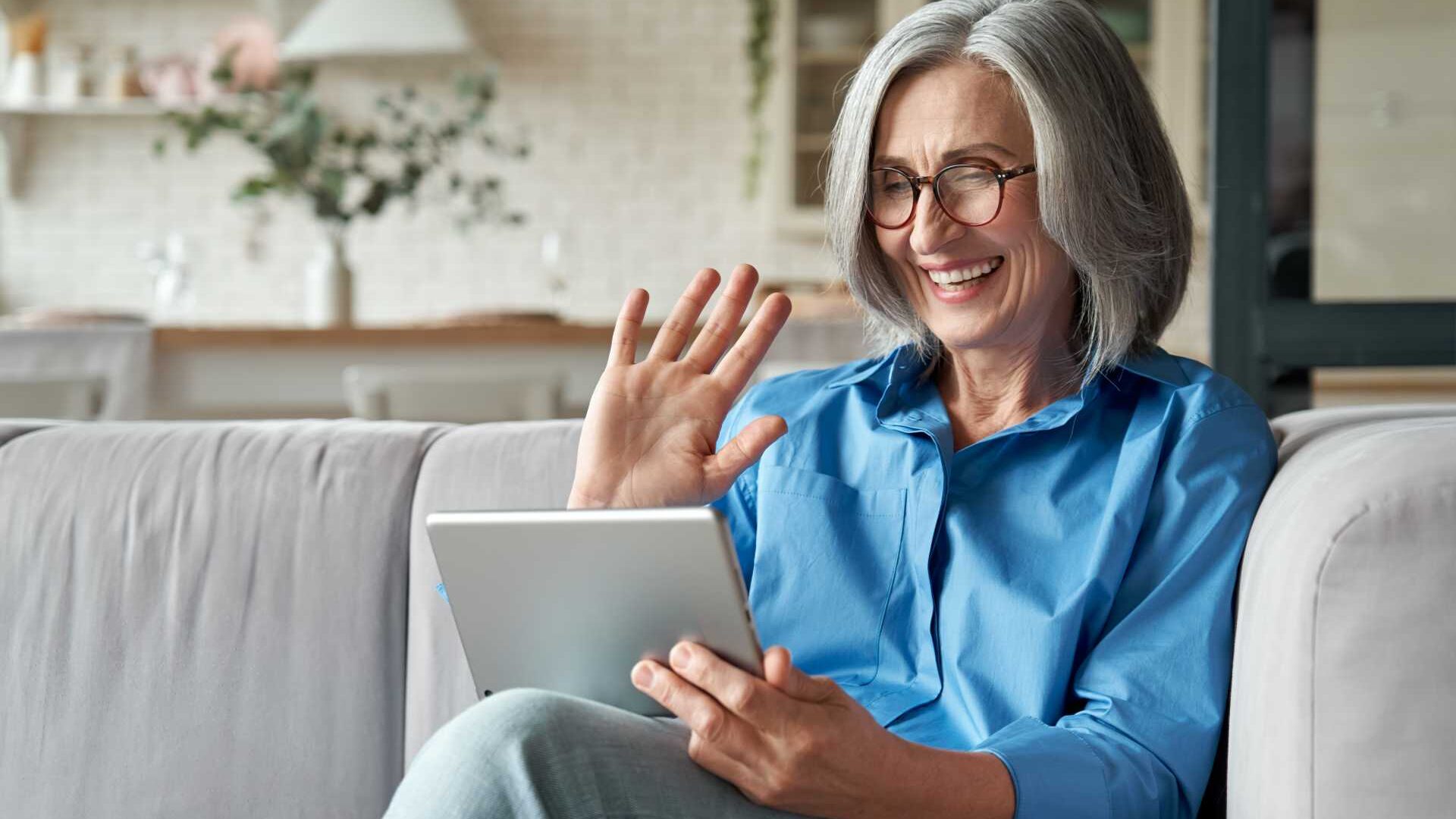 A woman waves on a virtual meeting while working from home, holding a tablet