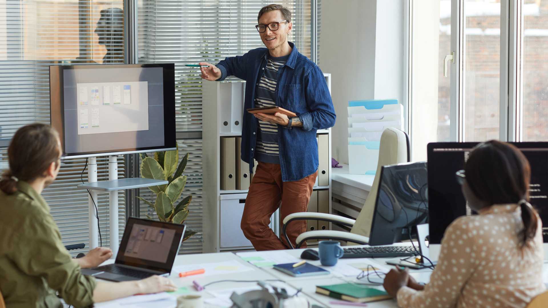 A salesperson is standing in front of employees as they look at different data figures on a monitor and laptop, representing empathy in sales management