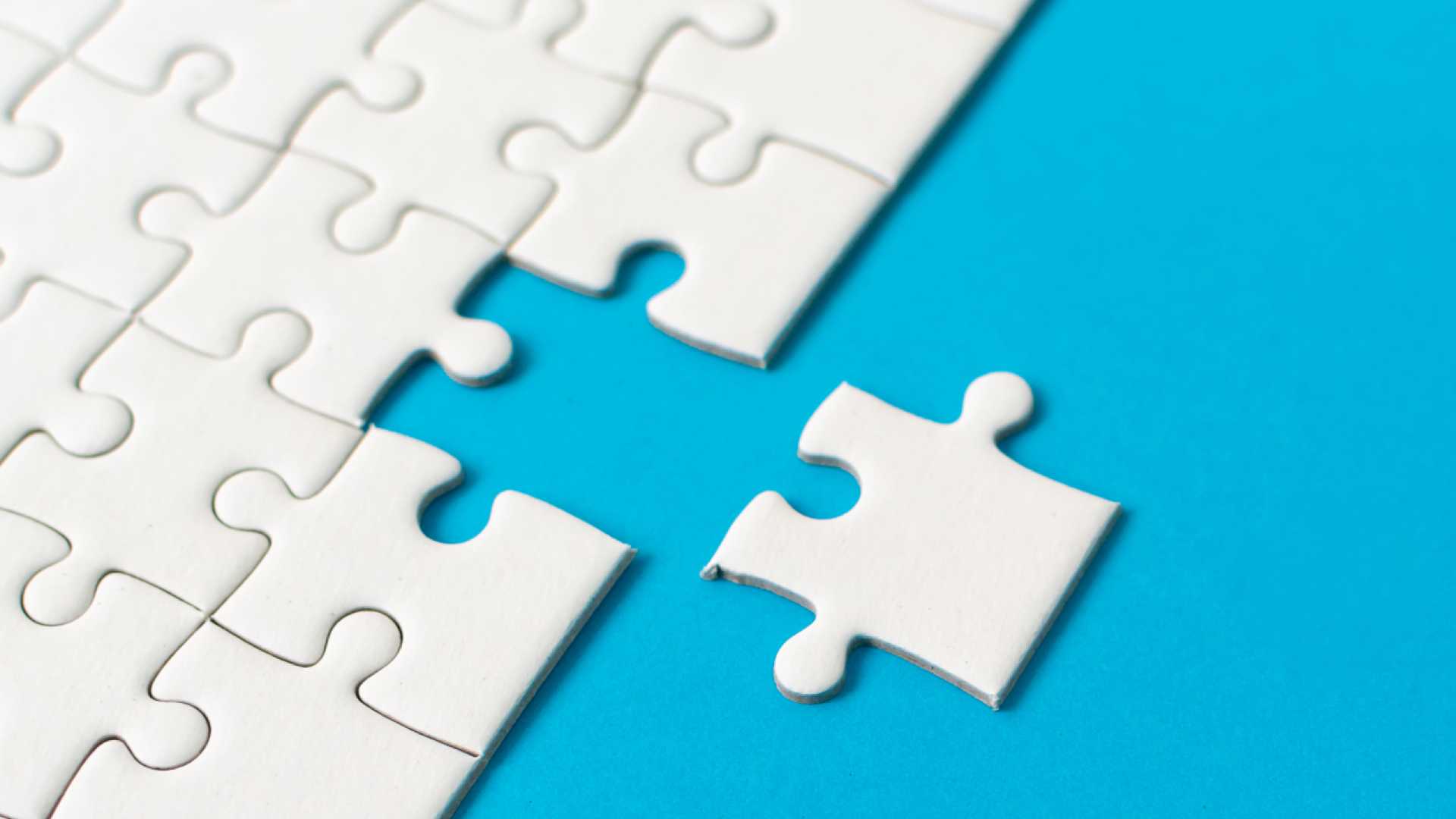 White puzzle piece fitting into an almost completed white jigsaw puzzle against a blue background.