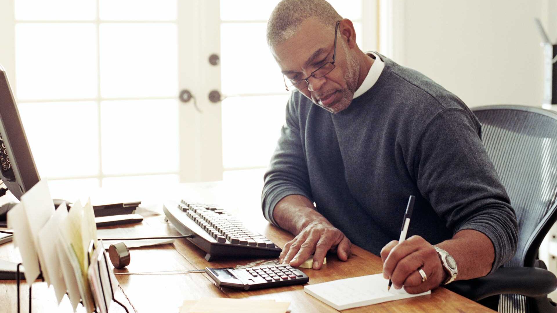 A man sitting at a desk in a home office space writing, on a notepad.