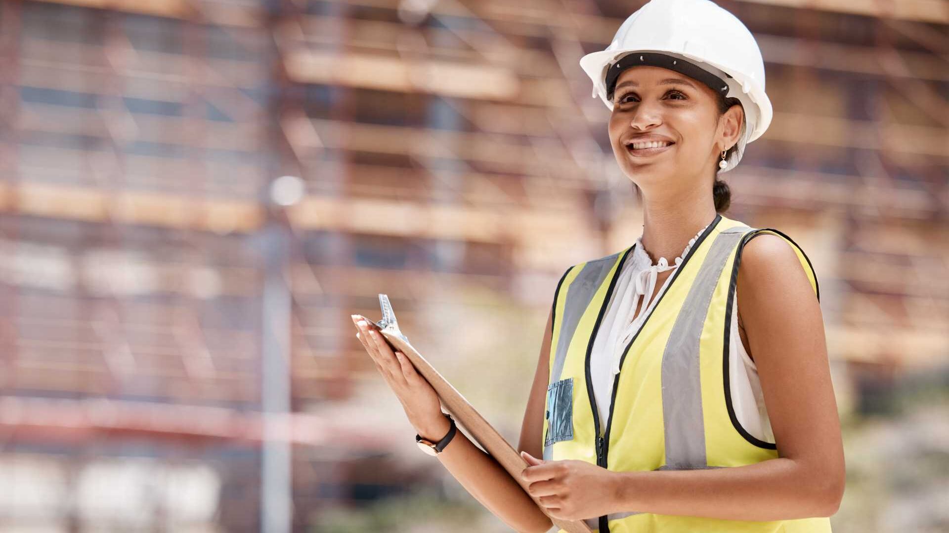 A woman wearing a construction hat and vest working in a manufacturing environment holding a clipboard