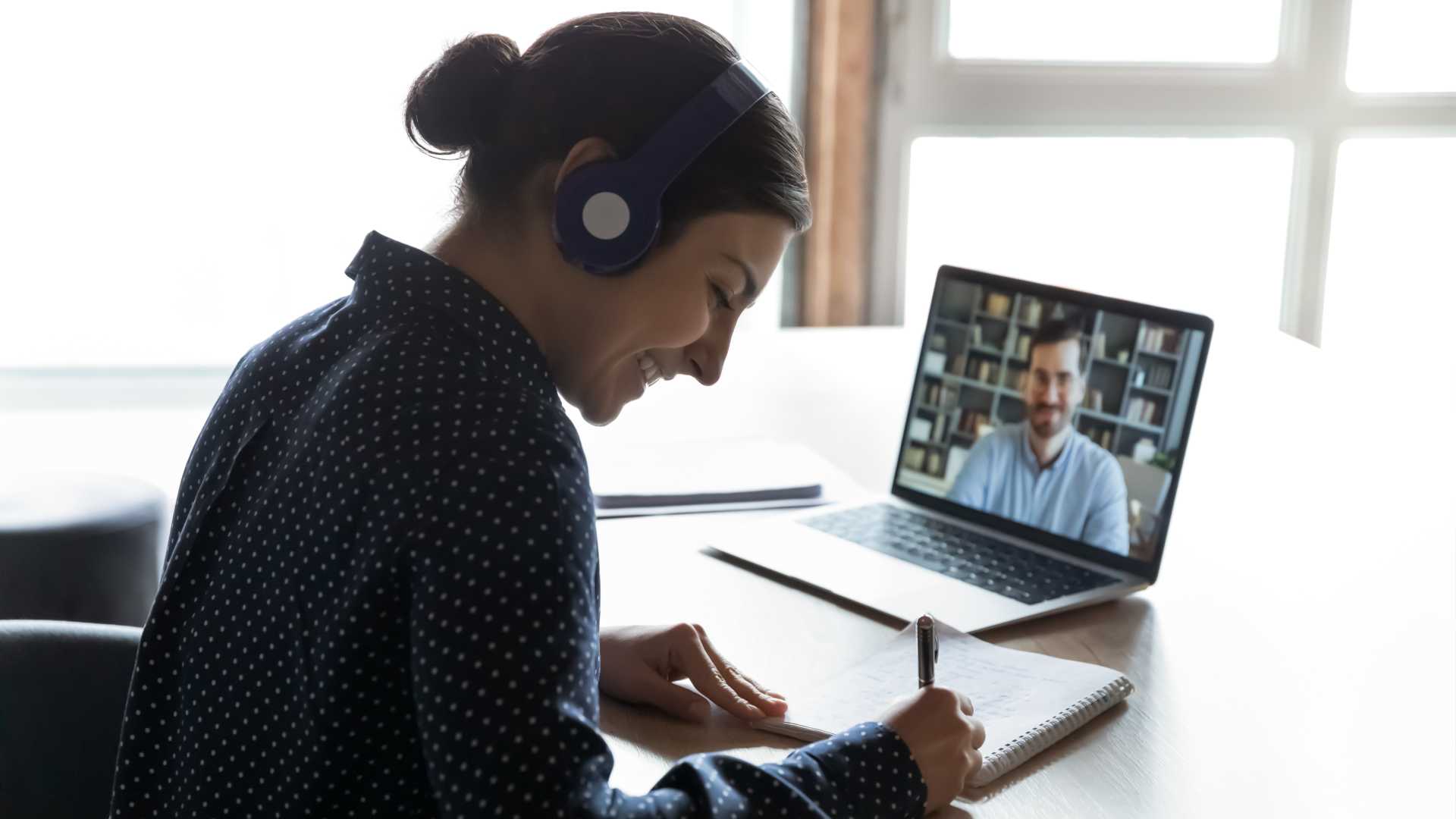 person with headphones on writing in a notebook while meeting with someone on a laptop