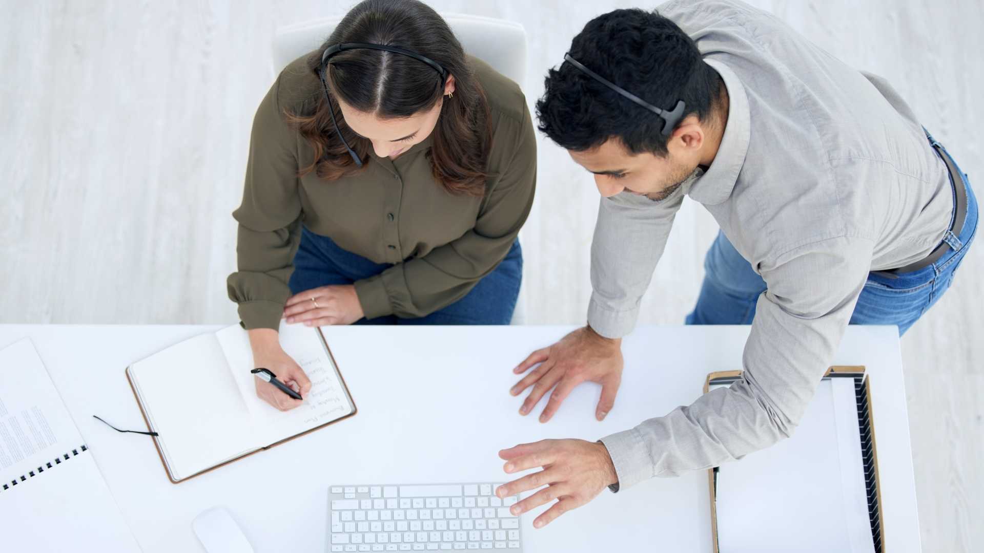 Two businesspeople collaborating in a work environment with one writing in a notebook and both wearing headsets