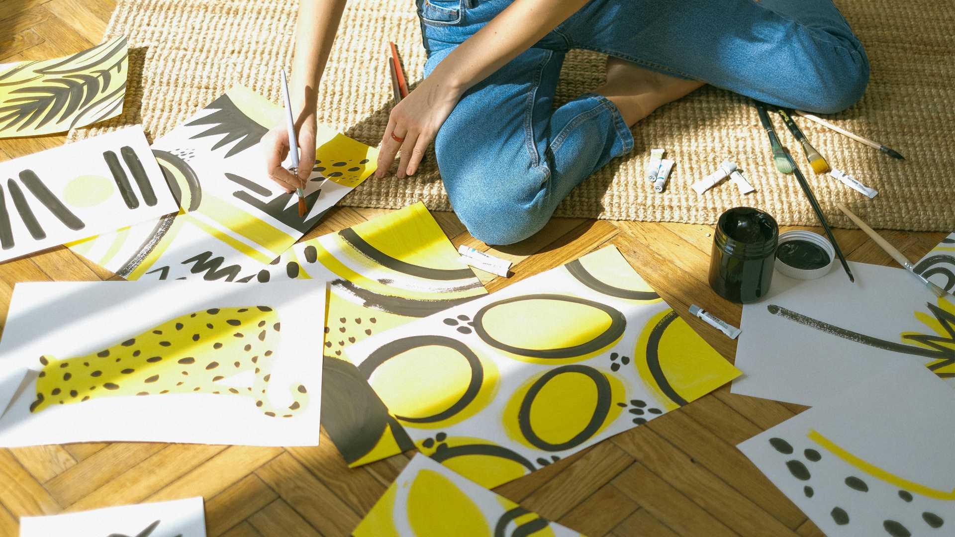 A woman seated on the floor holding a paintbrush surrounded by yellow abstract paintings