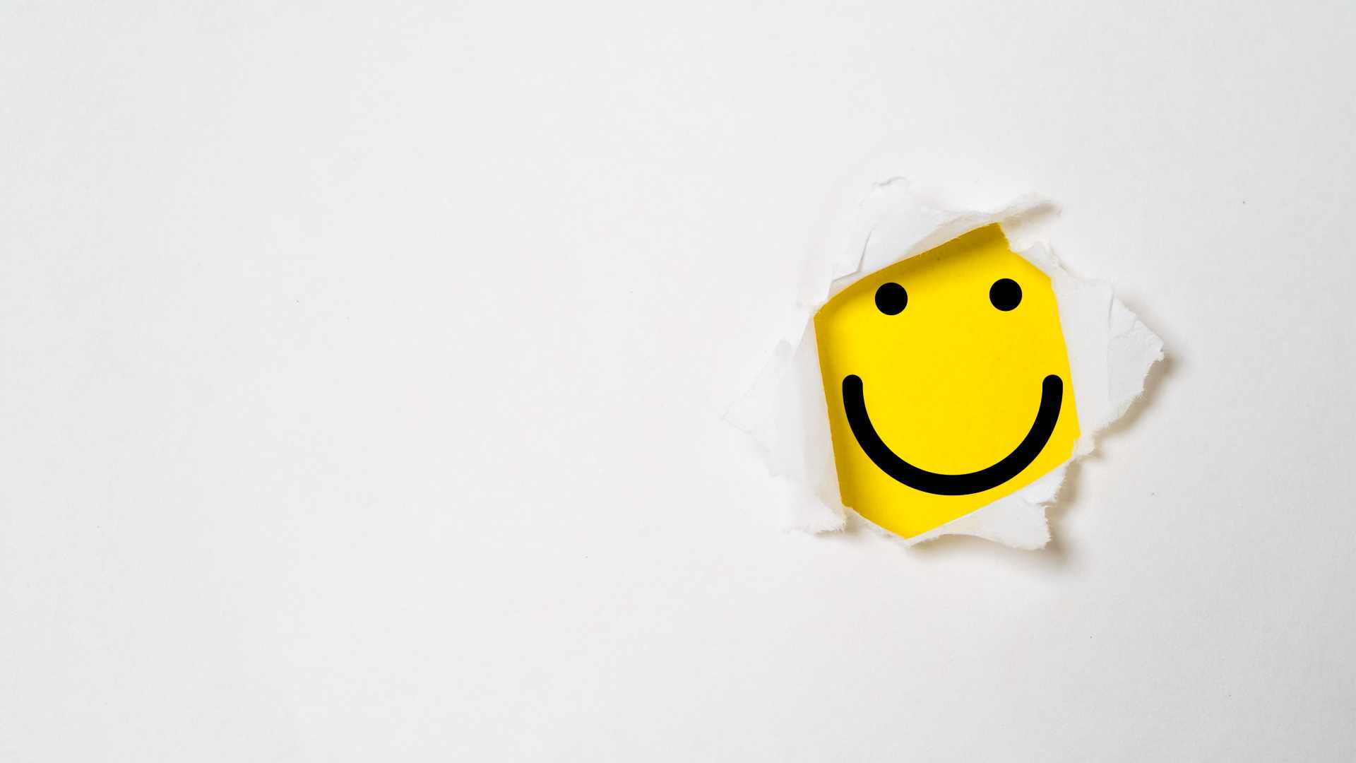 A yellow smiley face with two beady black dots for eyes and a long black inverted curve for a smile poking through a rip in white paper.