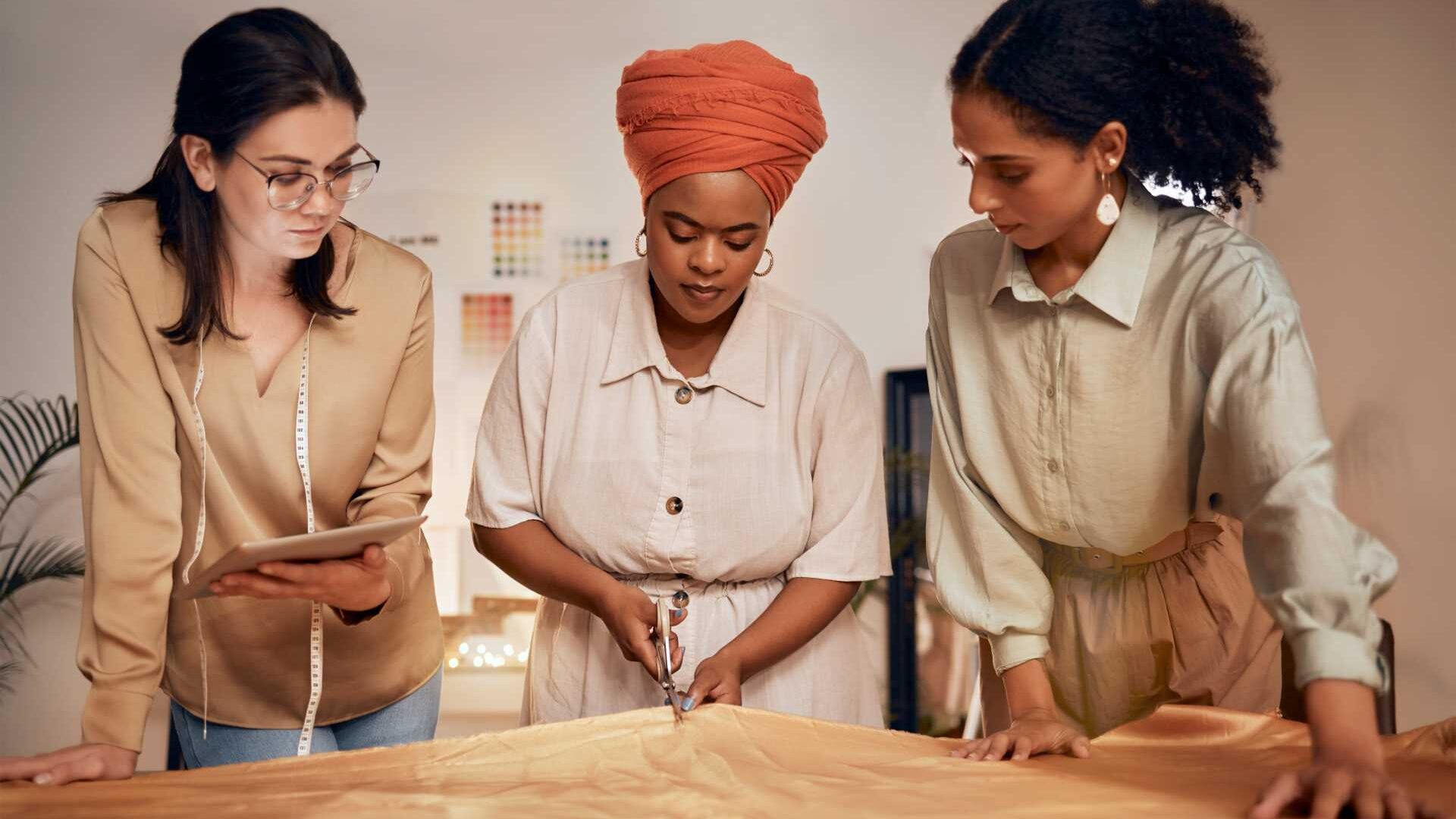Three females standing, one holding an iPad, one is examining fabric and one is cutting fabric.