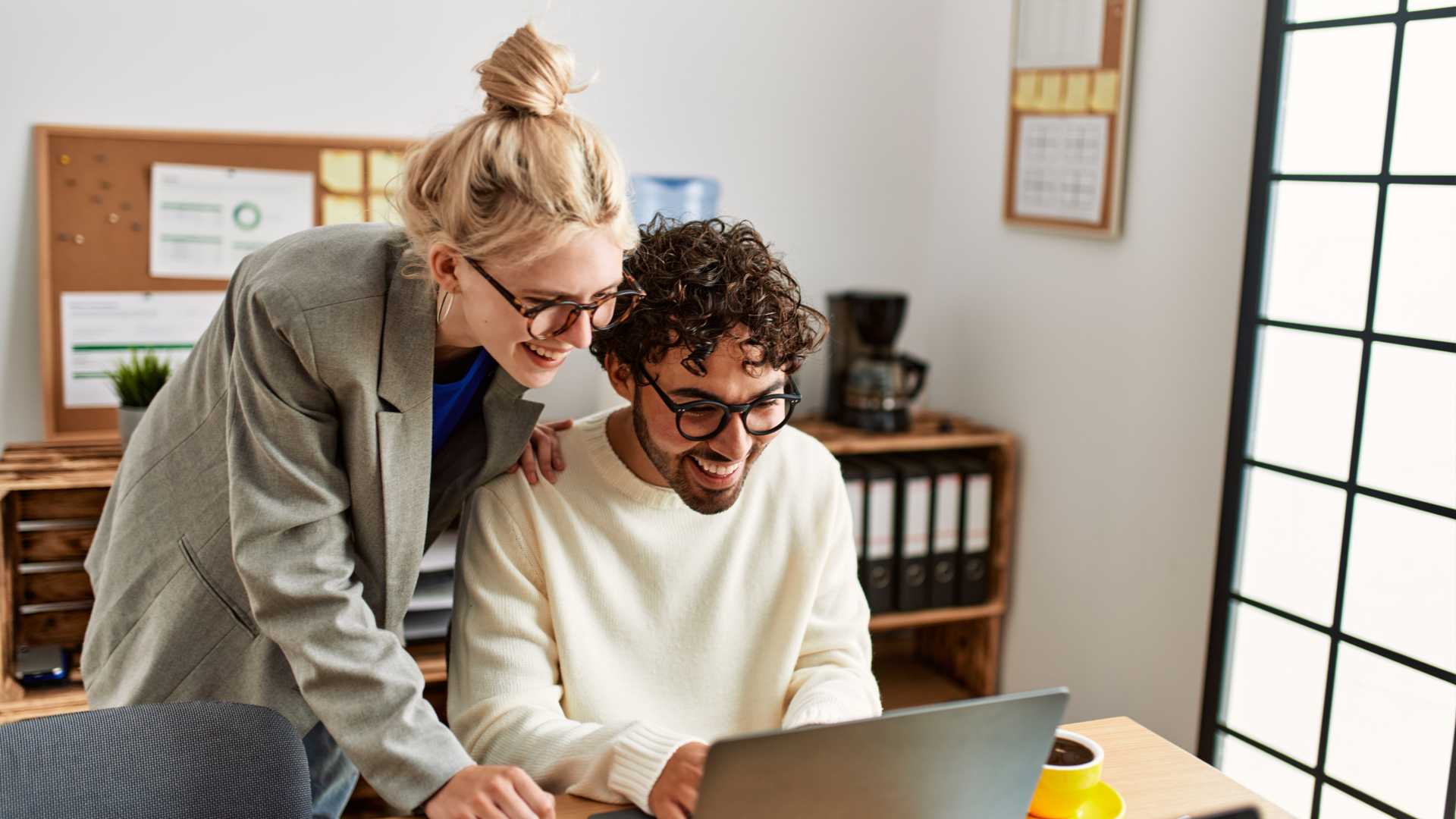 Two colleagues, one standing and one sitting, evaluating data on a laptop in a business corporate office environment.