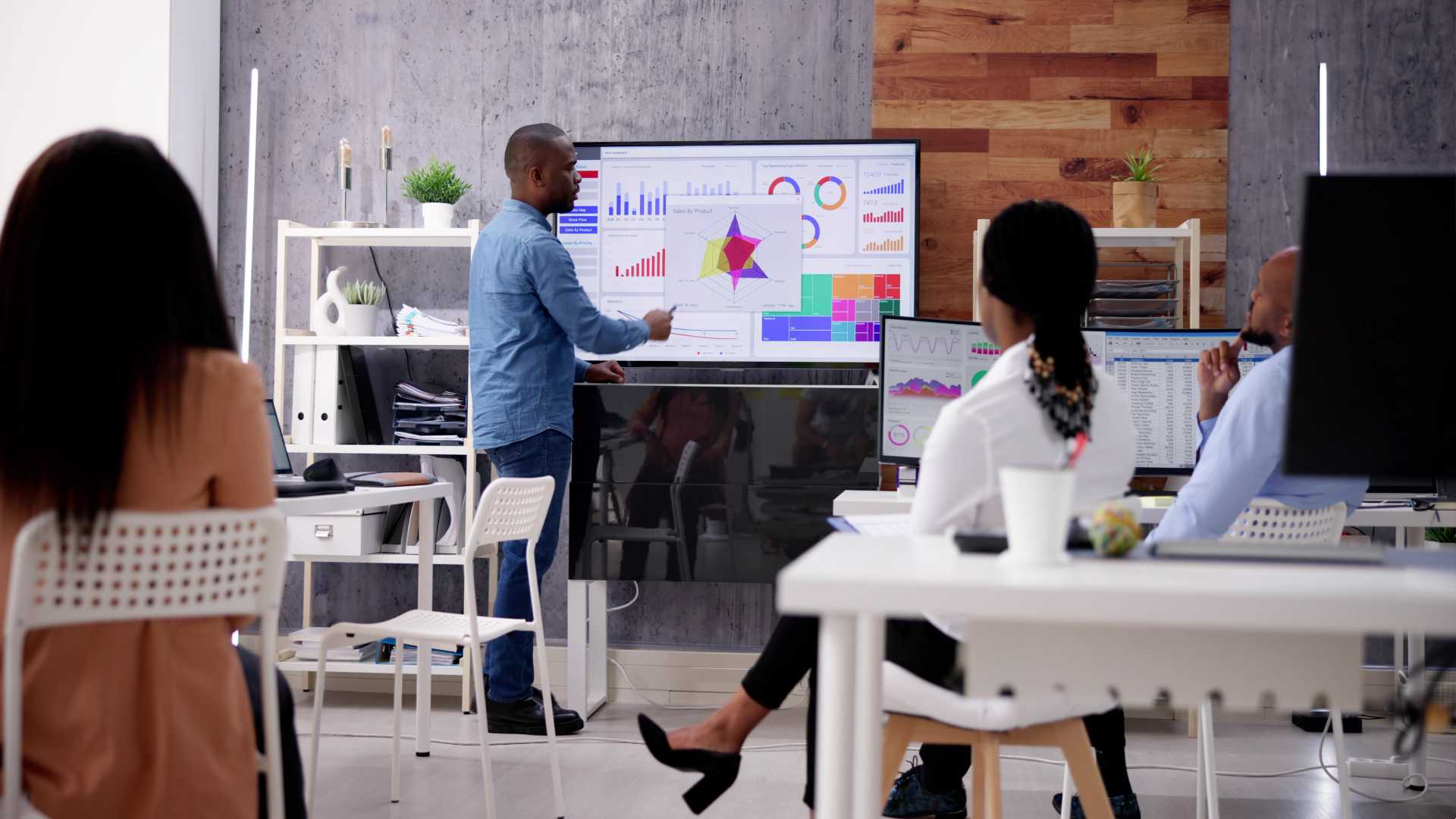 In an office setting, a man stands before a large screen while a group of colleagues observes his presentation.