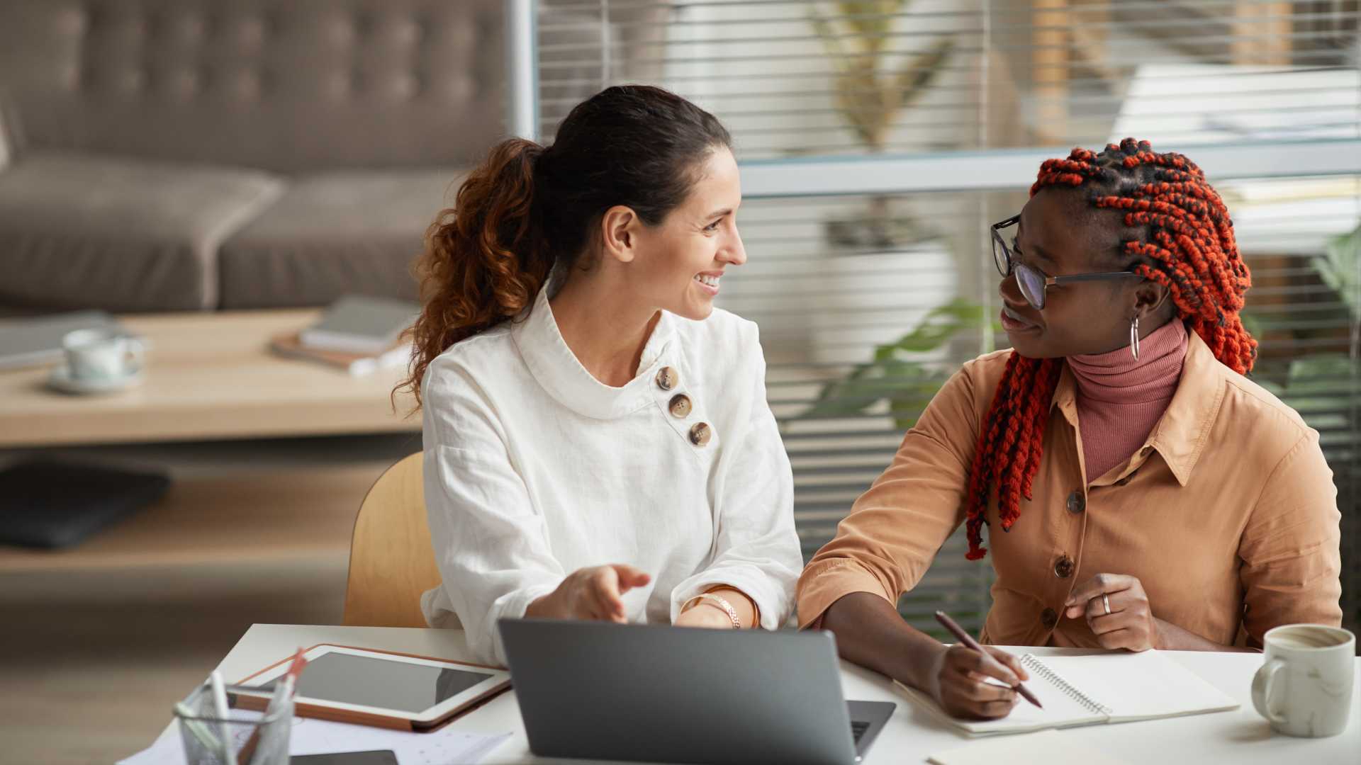 Portrait of two smiling businesswomen discussing project while sitting at desk together and working on startup in office.