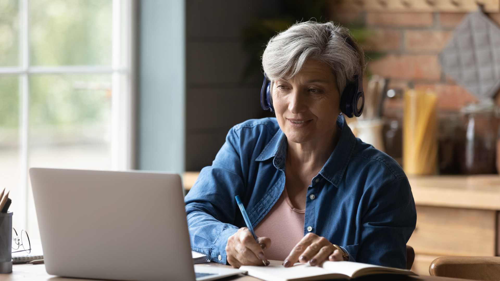 A woman seated in a work from home office looking at a computer and writing in a notebook
