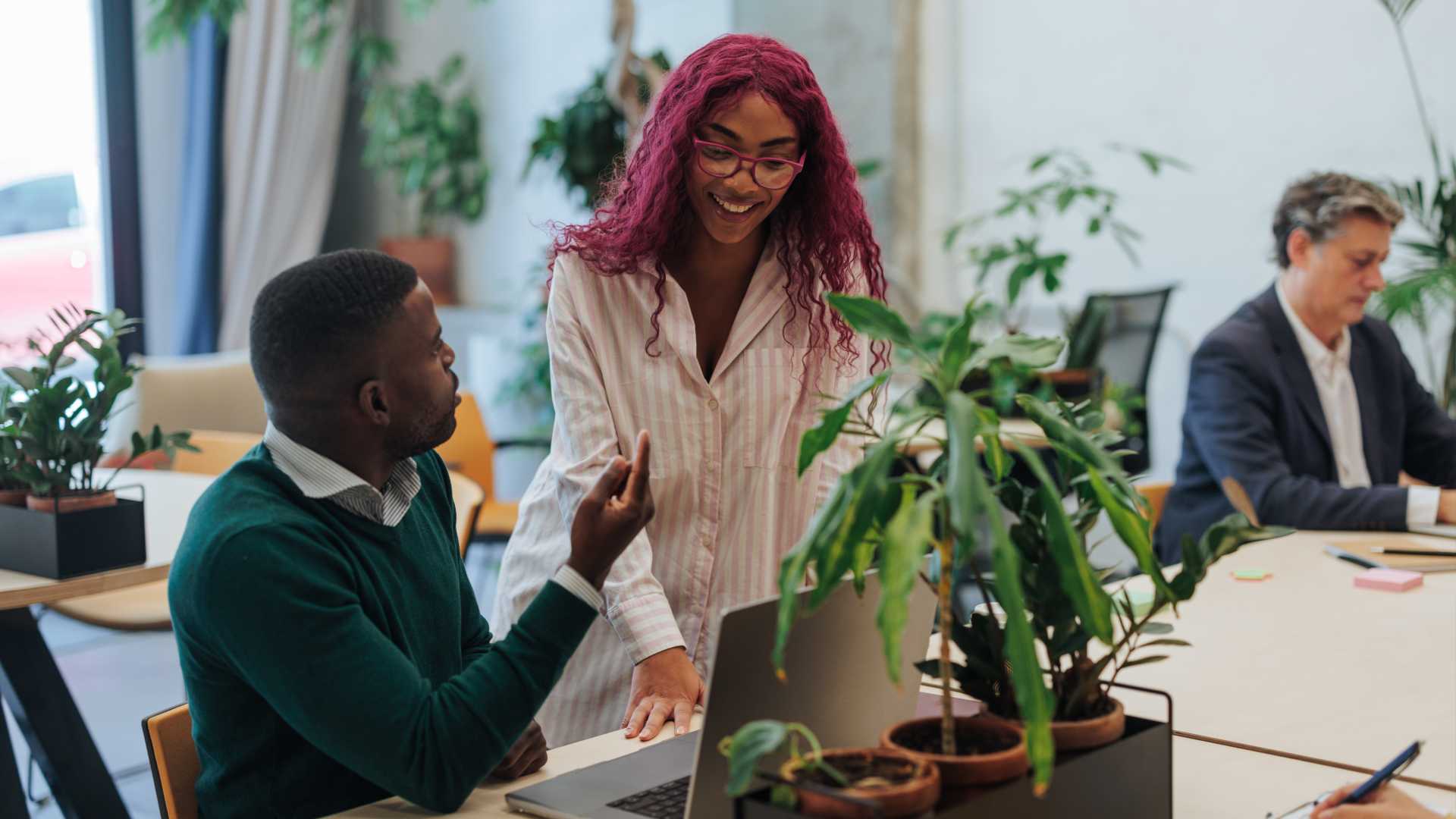 Two young professionals collaborating on a project, using a laptop in a modern, plant-filled office space, showcasing teamwork and a positive work environment