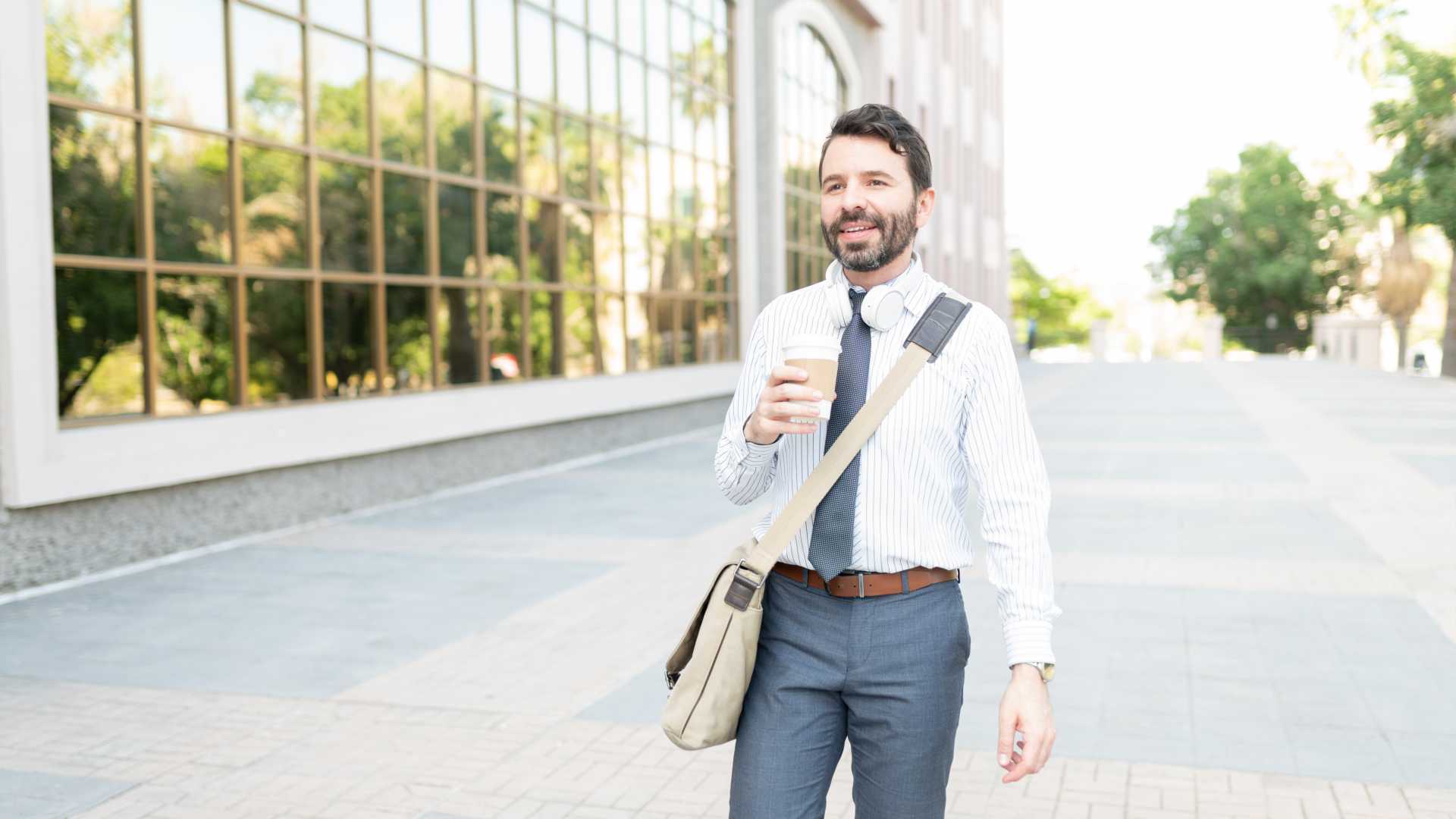 Business worker walking outside corporation with headphones around neck, satchel/cross body bag for work and holding a coffee cup.