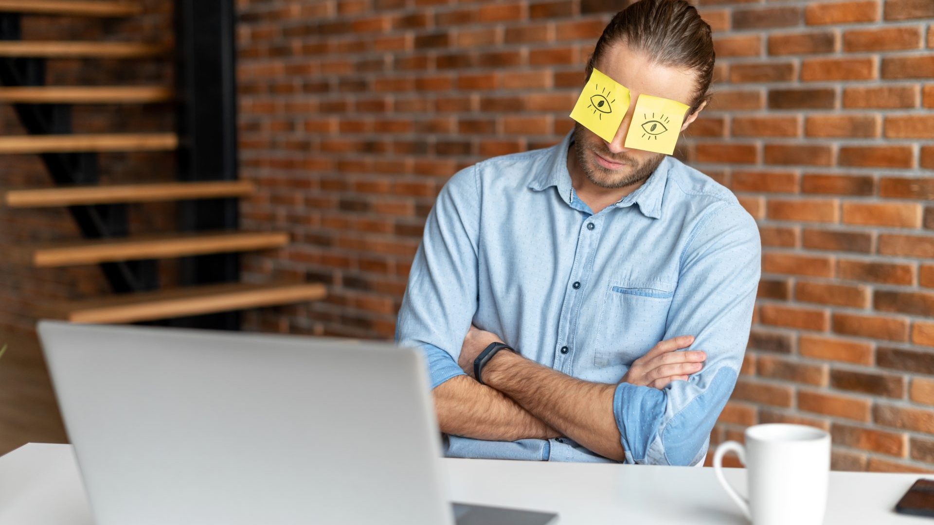 Man sits in front of an open laptop with arms crossed and post-it notes over his eyes.