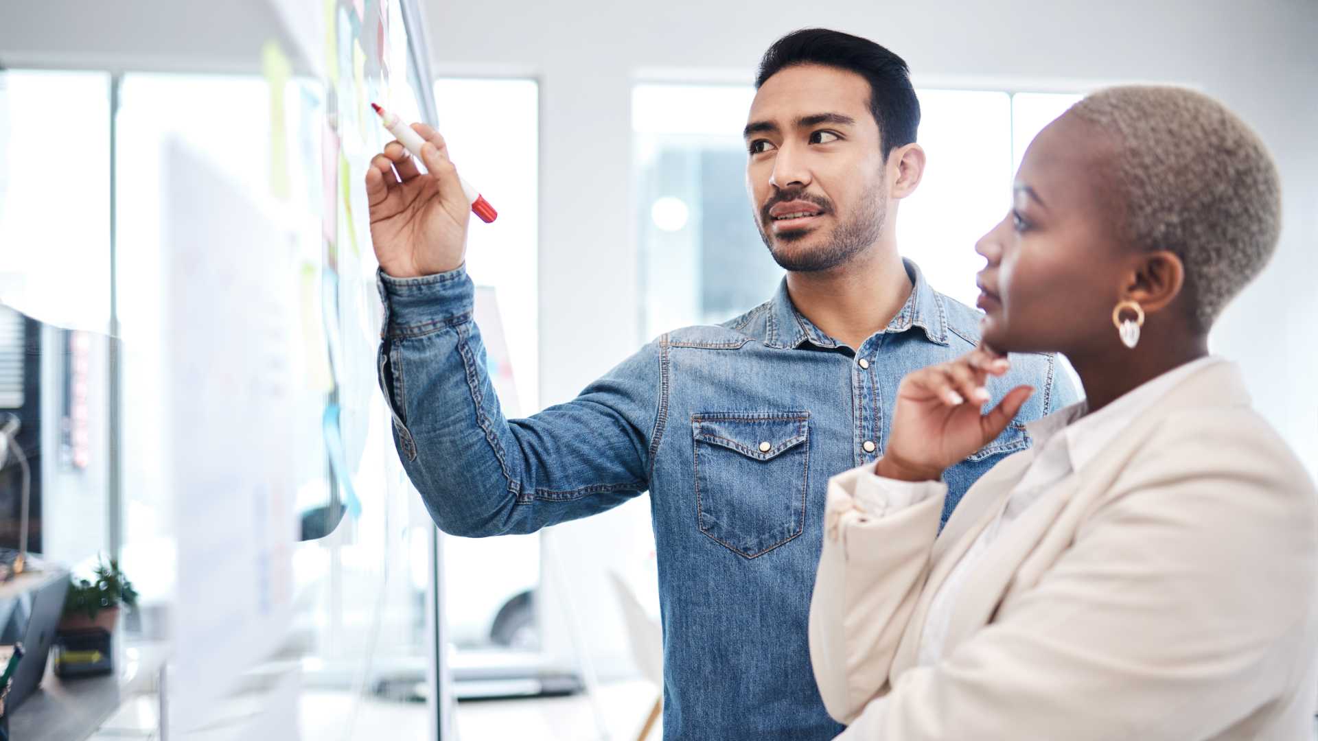 Male coworker teaching female coworker something and writing it on a whiteboard.