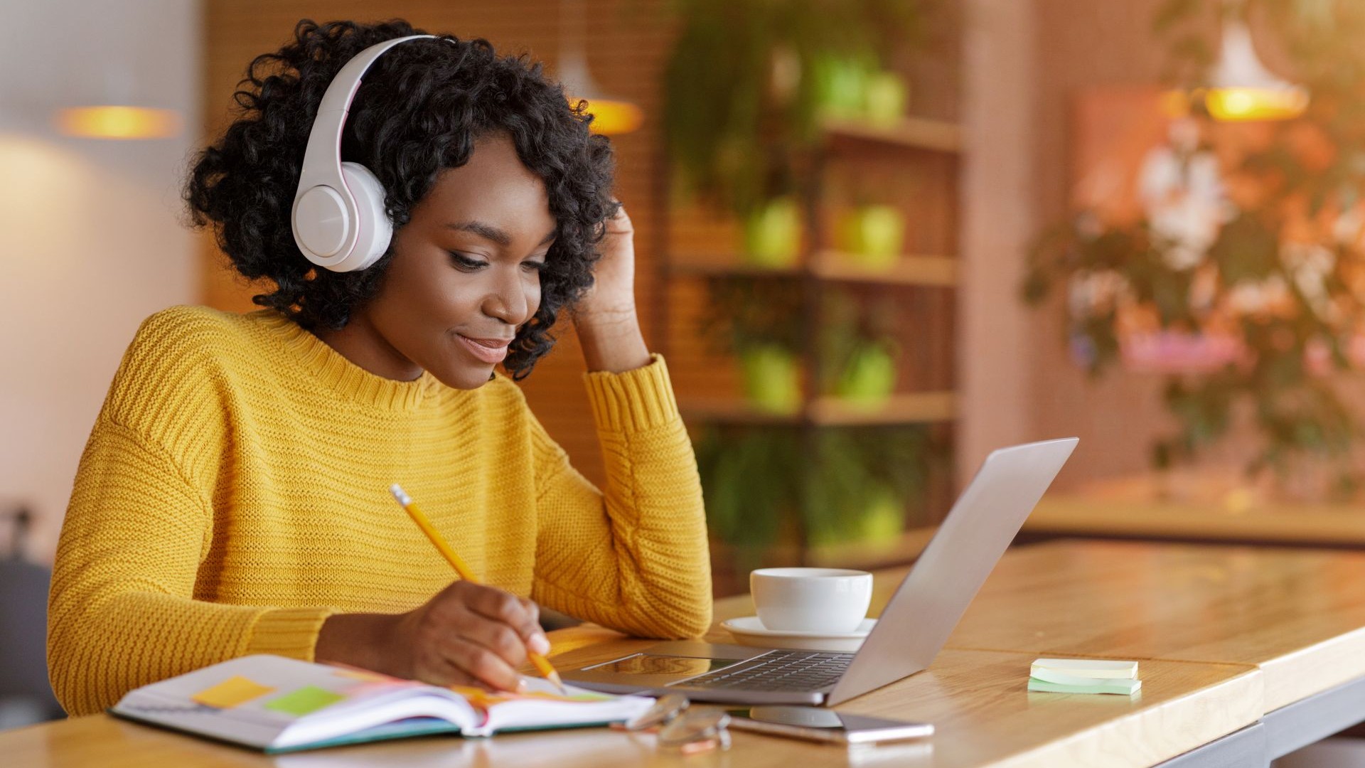 Woman in yellow sweater wearing headphones writes in a notebook while working on a laptop at a cozy café table.