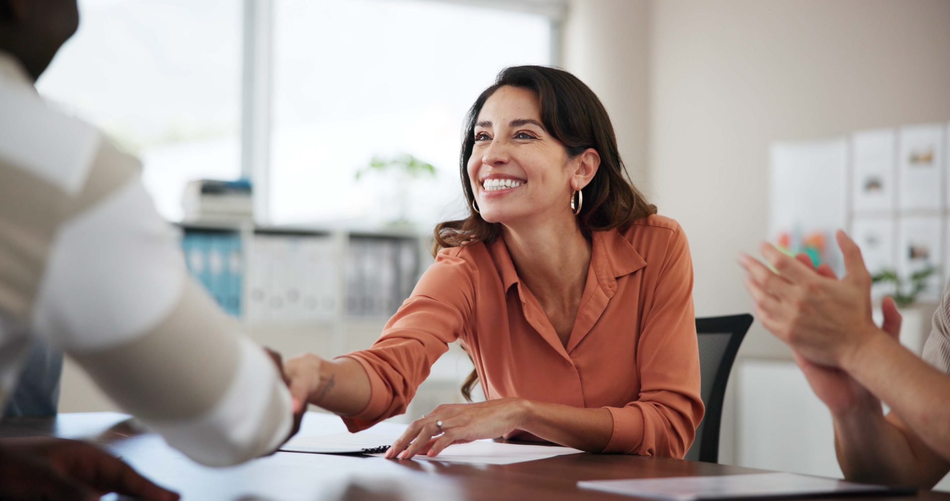 A woman sits at a desk in a professional setting. She shakes hands with another professional sitting across the desk. There are papers on the table. She looks happy.