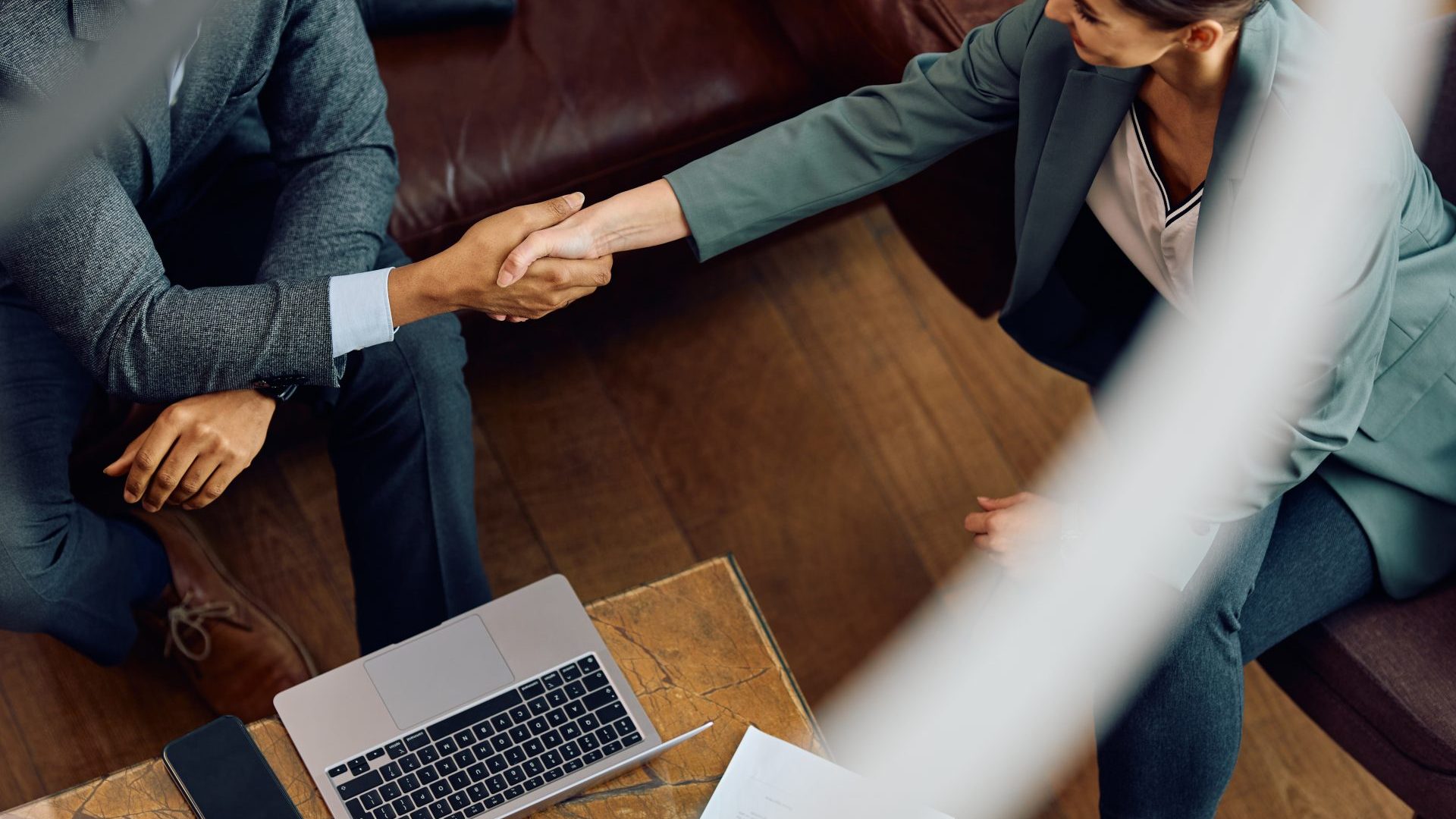 Overhead candid image of a woman shaking hands with a man. They are in a professional setting, with an open laptop on a desk between them.