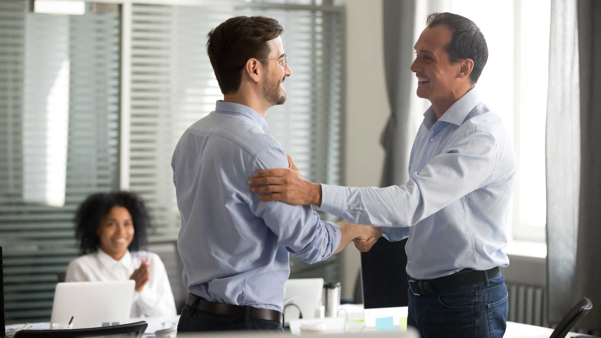 two business professionals shake hands and smile in an office while a colleague applauds in the background during a workplace recognition moment