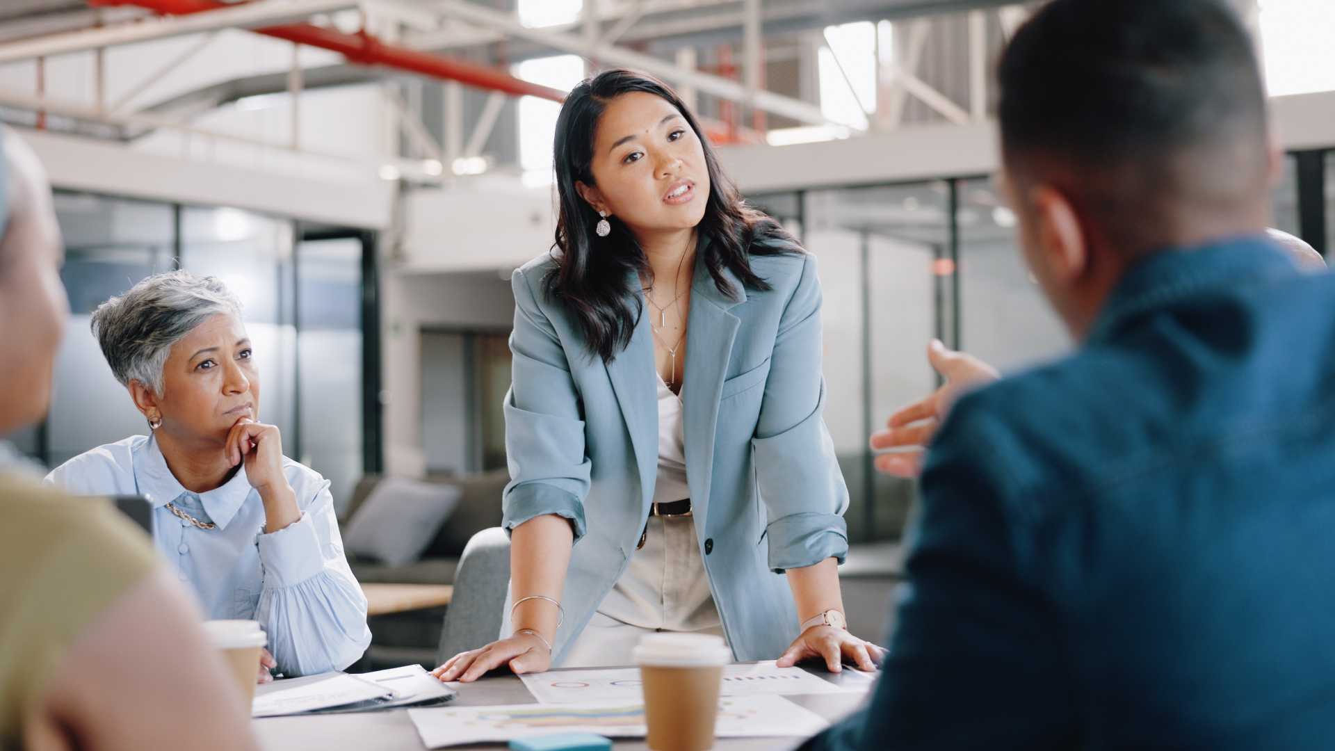 a business leader stands at a conference table speaking with colleagues during a team meeting while employees listen and participate in the discussion