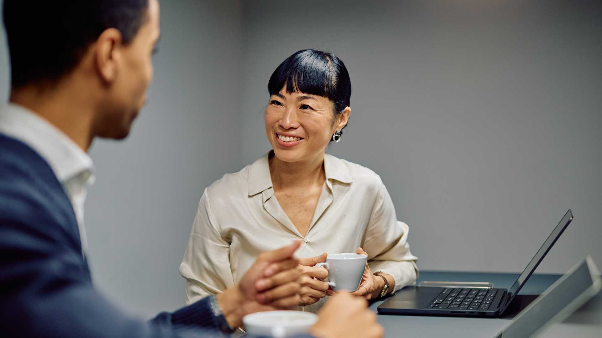 manager and employee having a one-on-one conversation over coffee at a table with an open laptop, illustrating supportive leadership, coaching and open workplace communication