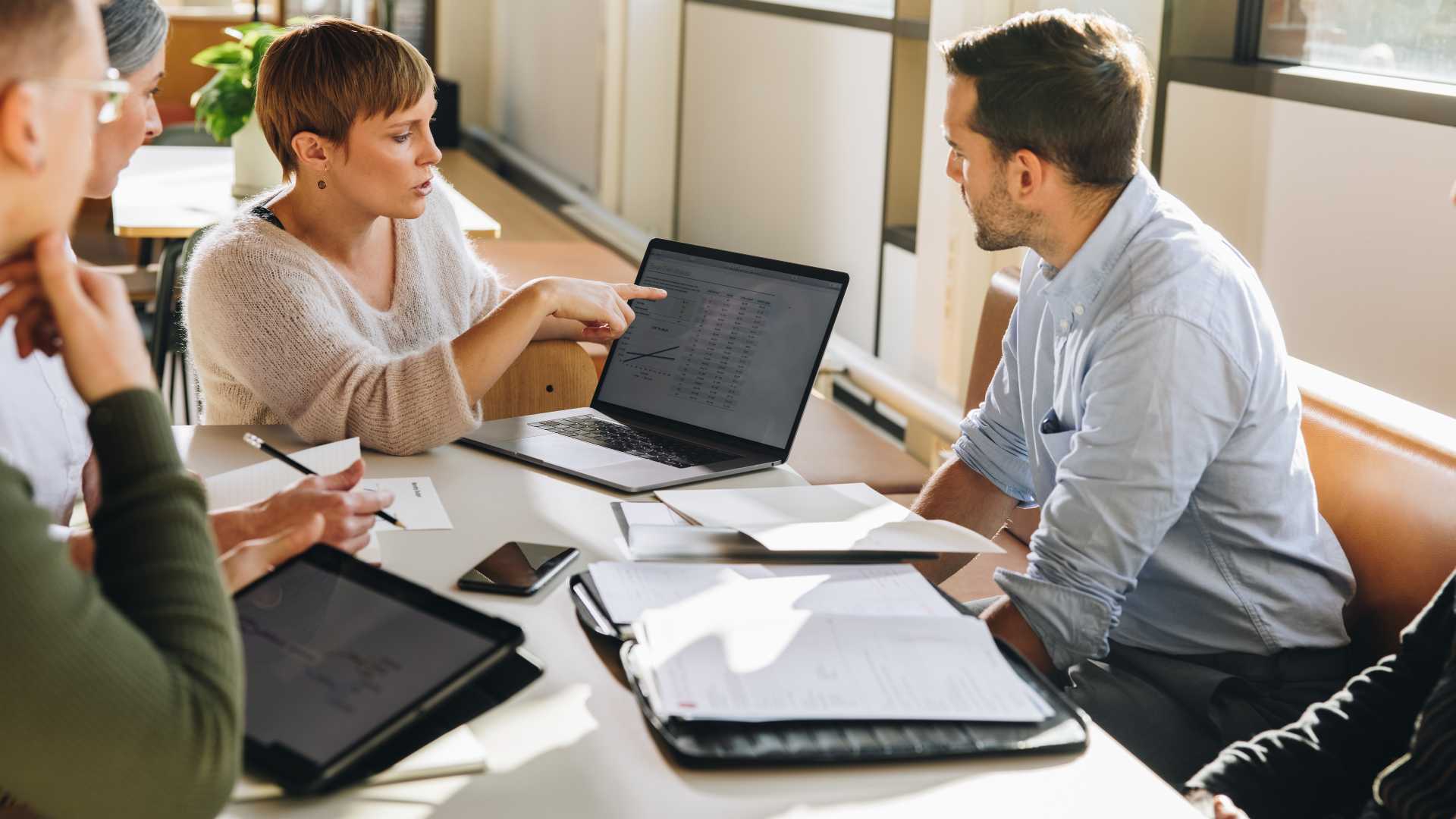 Business team discusses performance data and workplace priorities during a collaborative meeting with laptops and documents.