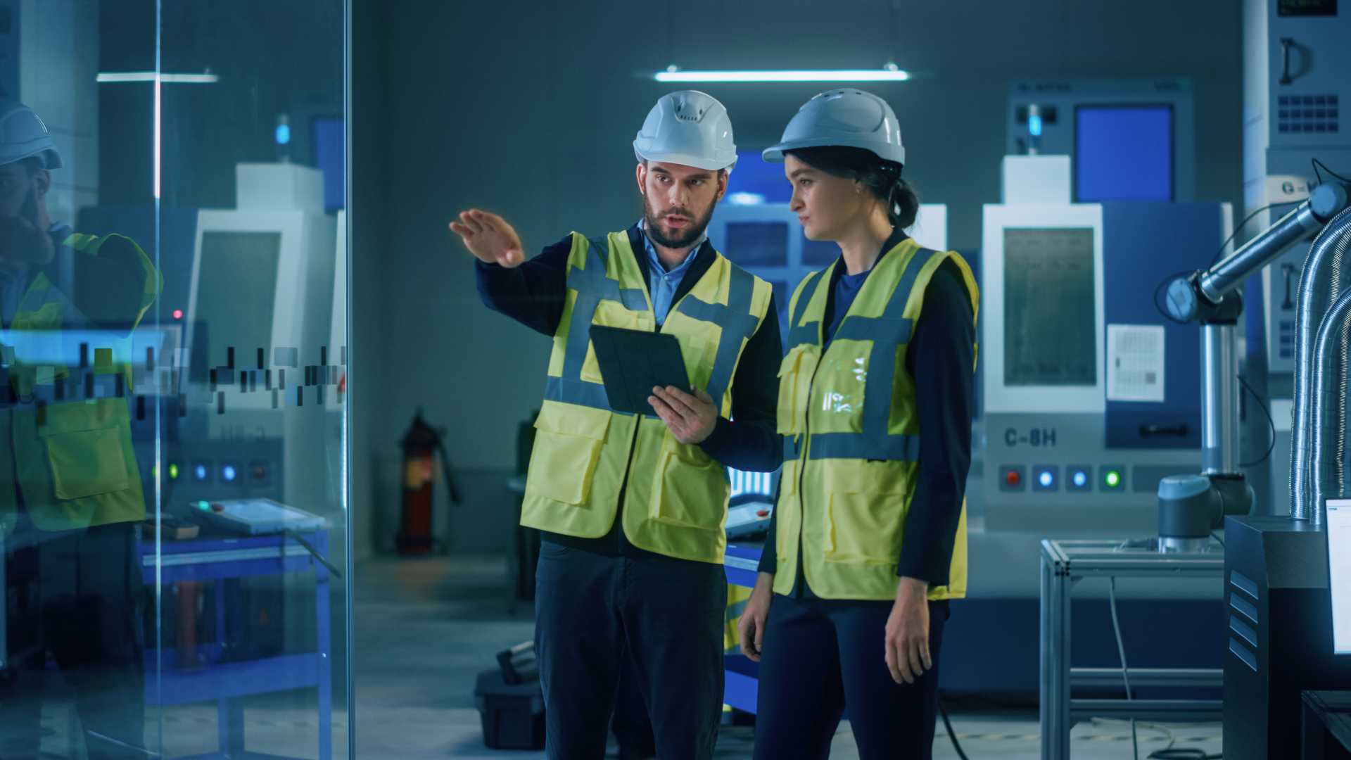 Two employees wearing hard hats and safety vests discuss equipment and safety procedures on a manufacturing floor.