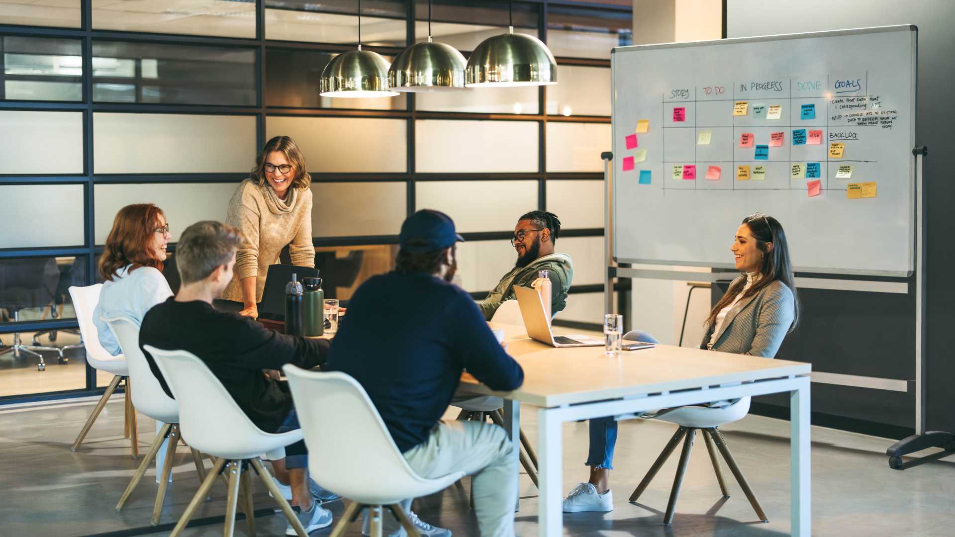 A manager leads a team discussion around a conference table with a project planning board in the background.