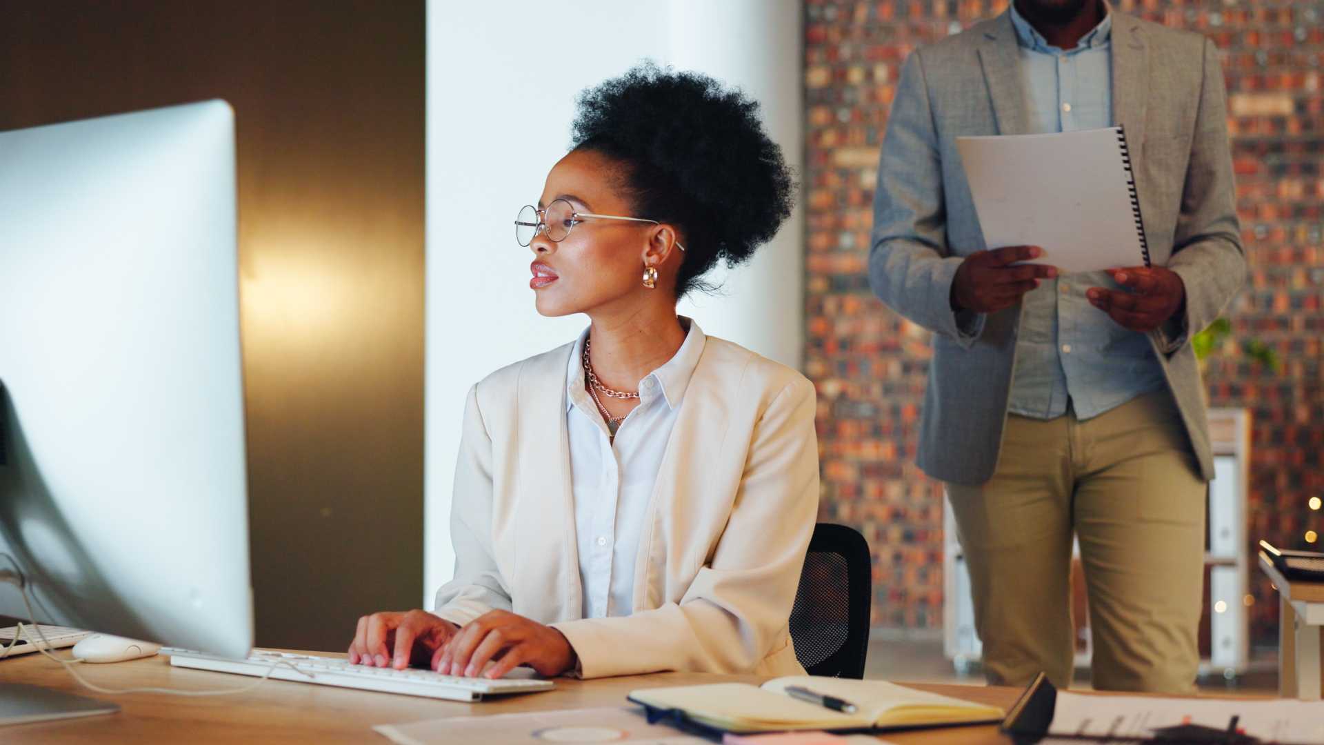 Employee working at a computer with a manager nearby, illustrating workplace coaching, performance expectations and accountability in a supportive leadership environment.