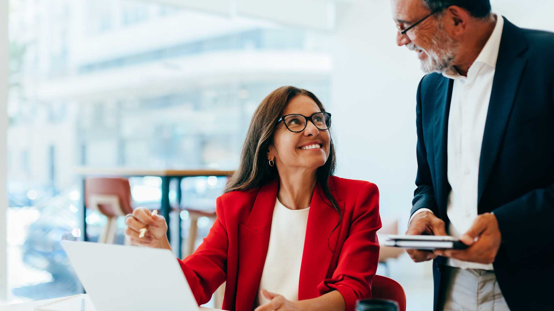 Businesswoman in a red blazer discussing work with a colleague beside a laptop, illustrating leadership coaching, collaboration and professional development.