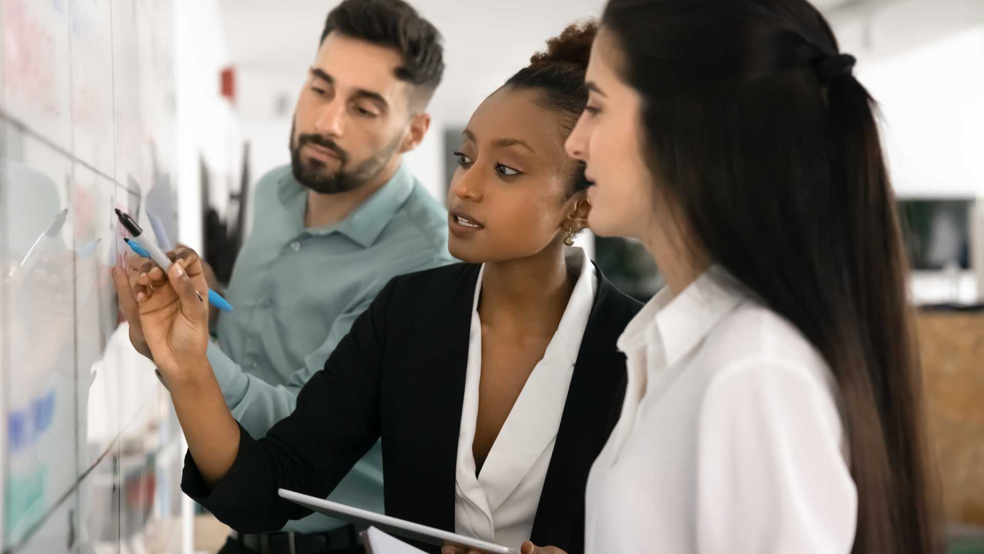 Three business professionals collaborating at a whiteboard, illustrating team problem-solving, decentralized decision-making and adaptive leadership in the workplace.